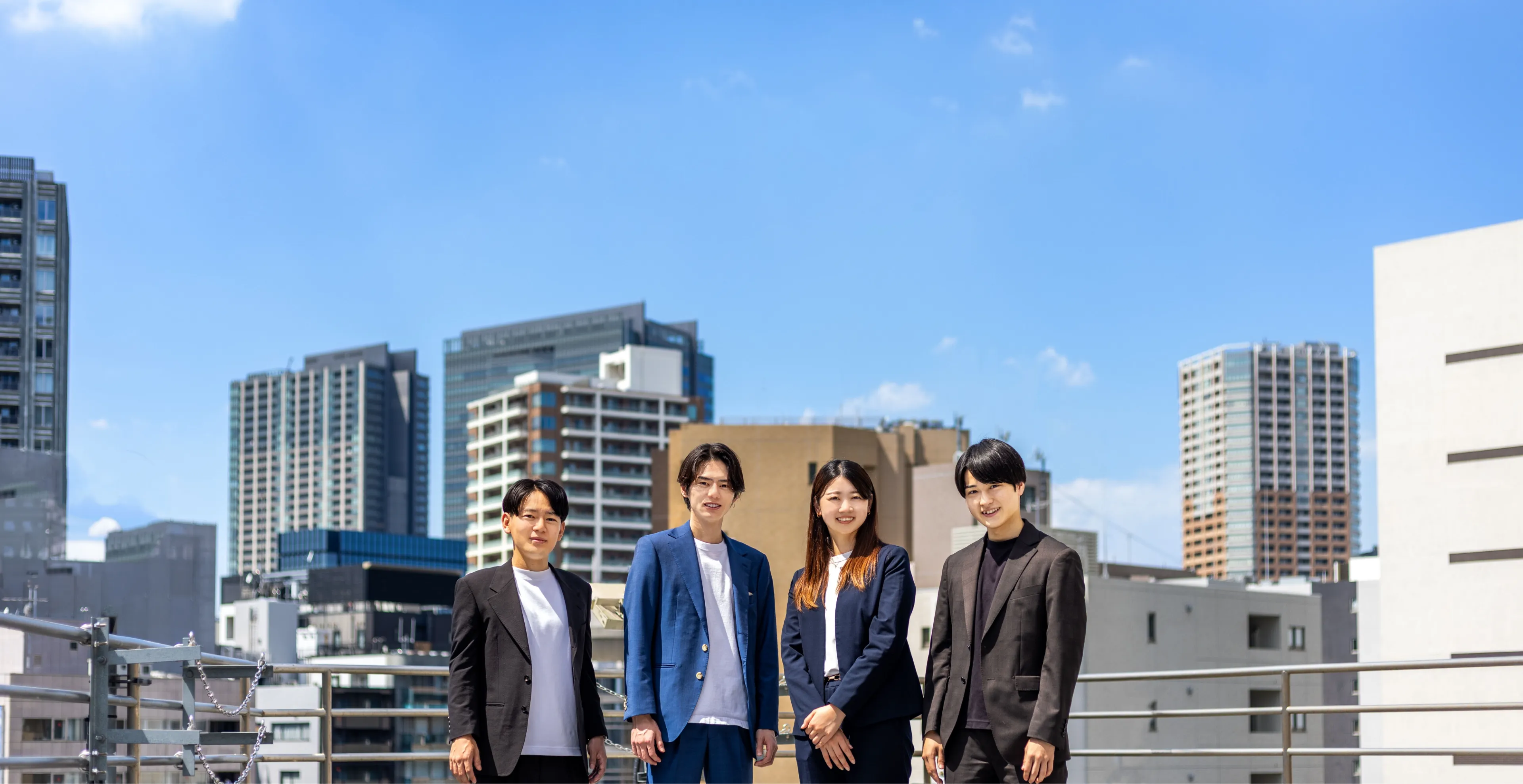 画像：ビル群を背景に男性3人、女性1人が並んで立っている様子