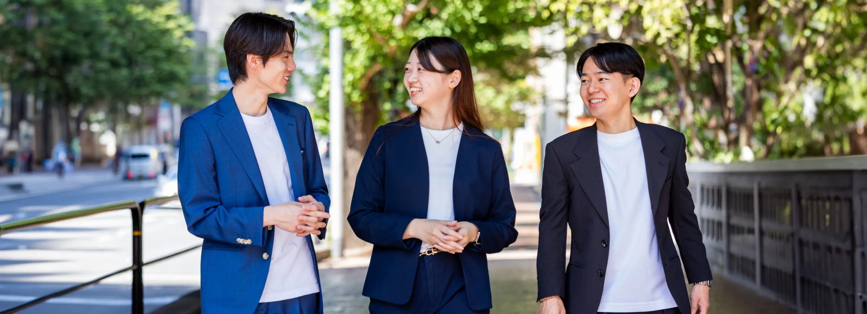 日差しの指す街中を、笑顔で談笑しながら歩く3人の男女社員。チームの仲の良さと活気を感じさせる風景